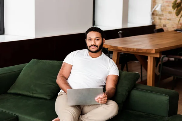 Barbuto afroamericano freelance seduto sul divano con computer portatile e guardando la fotocamera — Foto stock