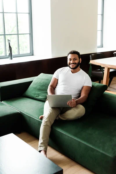 Happy african american freelancer sitting on sofa with laptop and looking at camera — Stock Photo