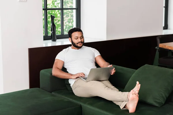 Beau freelance afro-américain avec pieds nus en utilisant un ordinateur portable dans le salon — Photo de stock