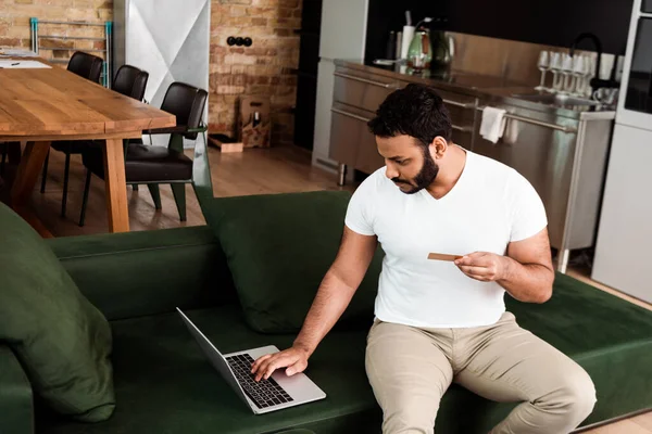 Barbudo hombre afroamericano con tarjeta de crédito cerca de la computadora portátil, mientras que las compras en línea en la sala de estar - foto de stock