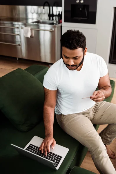 Hombre afroamericano sosteniendo la tarjeta de crédito cerca del ordenador portátil mientras que las compras en línea en la sala de estar - foto de stock
