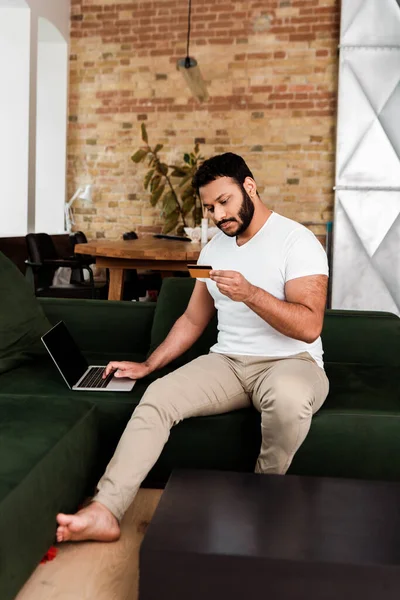 Hombre afroamericano que sostiene la tarjeta de crédito cerca del ordenador portátil con la pantalla en blanco mientras que compras en línea en la sala de estar - foto de stock