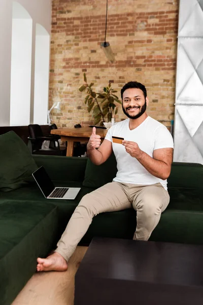 Fröhlicher afrikanisch-amerikanischer Mann hält Kreditkarte neben Laptop mit leerem Bildschirm, während er Daumen nach oben zeigt — Stockfoto