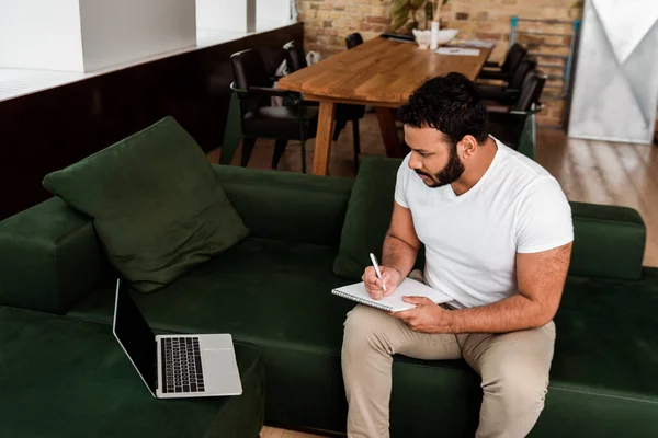 Bärtiger afrikanisch-amerikanischer Mann schreibt in Notizblock in der Nähe von Laptop mit leerem Bildschirm, Studie Online-Konzept — Stockfoto