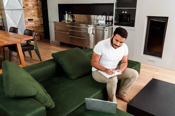 Schöner afrikanisch-amerikanischer Mann schreibt in Notizbuch in der Nähe von Laptop, Studie Online-Konzept — Stockfoto