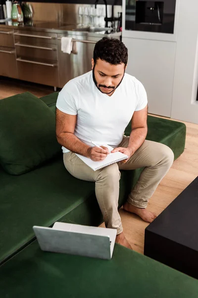 Бородатый африканский американец пишет в блокноте возле ноутбука, изучает онлайн концепцию — стоковое фото