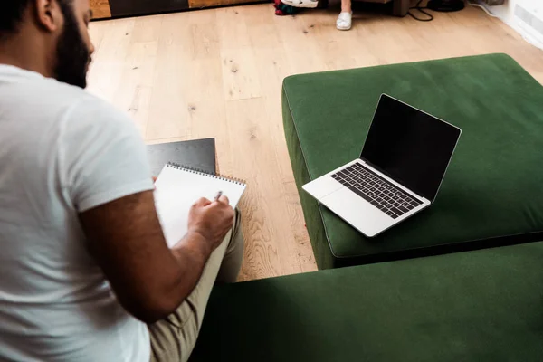 Selective focus of bearded african american man writing in notebook near laptop with blank screen, study online concept — Stock Photo