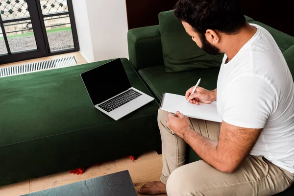 Barbuto uomo africano americano che scrive in notebook vicino al computer portatile con schermo bianco, studio concetto online — Foto stock