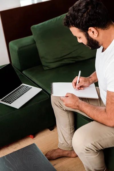 Barbuto uomo africano americano che scrive in notebook vicino al computer portatile con schermo bianco, studio concetto online — Foto stock