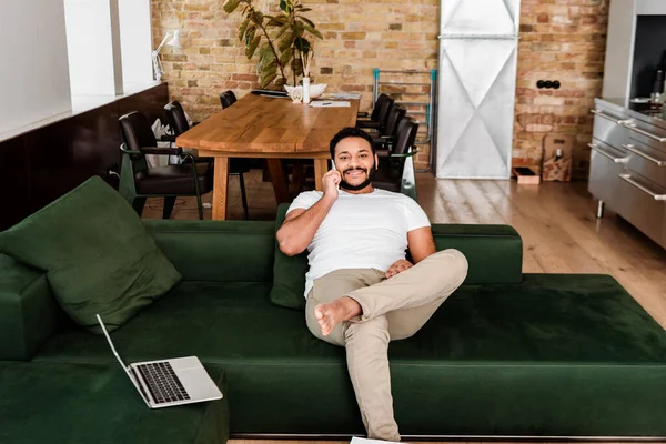 Щасливий афроамериканський чоловік говорить на смартфоні біля ноутбука на дивані — стокове фото