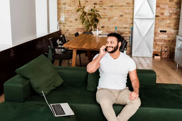 Alegre afroamericano freelancer hablando en smartphone cerca del portátil en el sofá - foto de stock