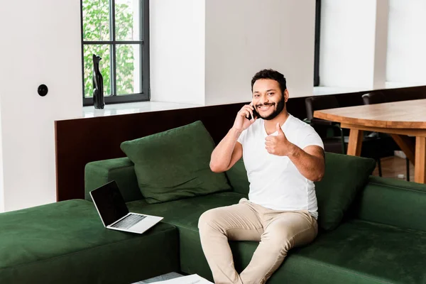 Homem americano africano barbudo falando no smartphone e mostrando o polegar perto do laptop com tela em branco — Fotografia de Stock