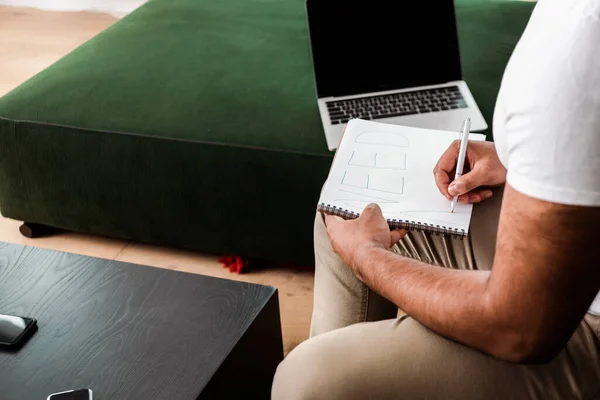 Abgeschnittene Ansicht eines Mannes, der ein Wort in ein Notizbuch in der Nähe eines Laptops mit leerem Bildschirm schreibt — Stockfoto