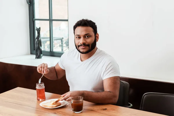 Barbuto uomo africano americano tenendo cucchiaio vicino vaso con marmellata e pane tostato sul piatto — Foto stock