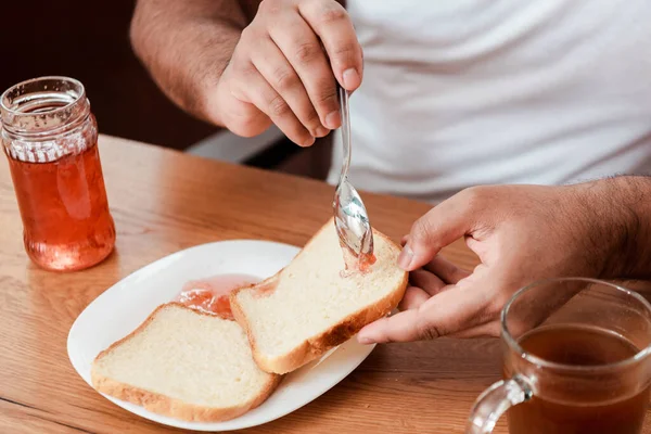 Vista recortada del hombre afroamericano sosteniendo cuchara con mermelada dulce cerca de pan tostado en el plato - foto de stock