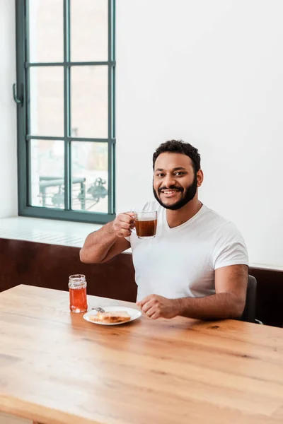 Sonriente afroamericano hombre sosteniendo taza de café cerca de pan tostado con mermelada dulce - foto de stock