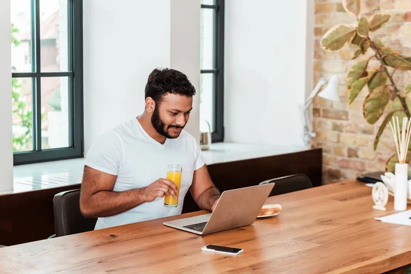 Freelancer americano africano barbudo trabalhando em casa e segurando um copo de suco de laranja — Fotografia de Stock