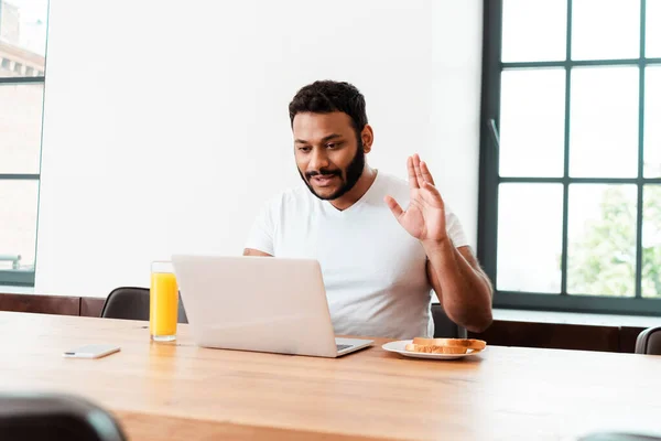 Selektiver Fokus eines bärtigen afrikanisch-amerikanischen Mannes, der beim Videoanruf in der Nähe eines leckeren Frühstücks auf dem Tisch winkt — Stockfoto