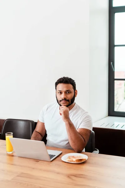 Barbudo afroamericano freelancer mirando a la cámara cerca del ordenador portátil y desayuno en la mesa - foto de stock