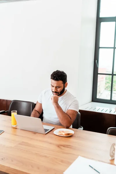 Pensoso africano americano freelancer guardando computer portatile vicino a colazione sul tavolo — Foto stock