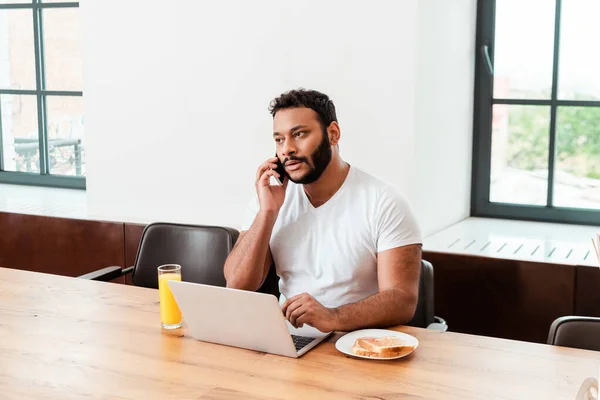 Freelancer afro-americano falando no smartphone perto de laptop e café da manhã na mesa — Fotografia de Stock