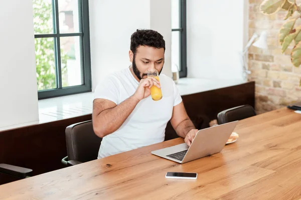 African american freelancer drinking fresh orange juice while using laptop near smartphone with blank screen — Stock Photo