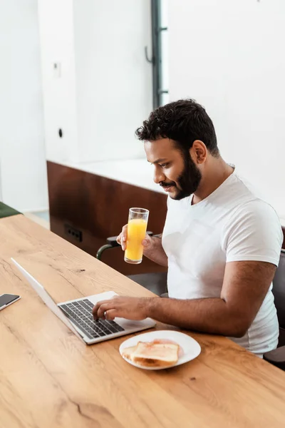 African american freelancer holding glass of fresh orange juice while using laptop near smartphone with blank screen — Stock Photo