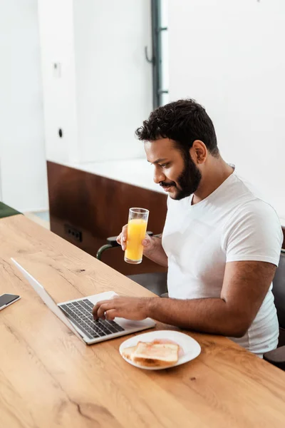 Afrikanisch-amerikanischer Freiberufler hält ein Glas frischen Orangensaft in der Hand, während er Laptop neben Smartphone mit leerem Bildschirm benutzt — Stockfoto