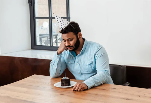 Sad african american man in party cap celebrating birthday alone and looking at birthday cake — Stock Photo