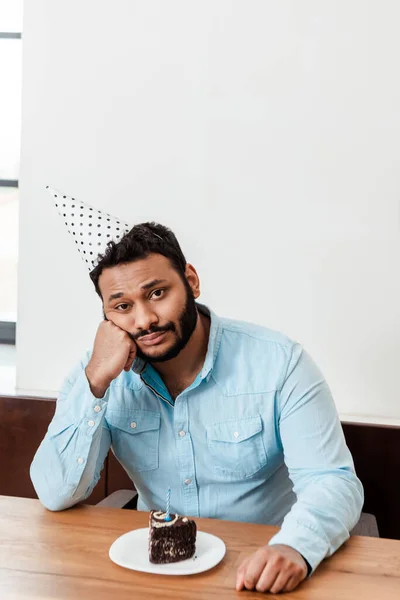 Upset african american man in party cap celebrating birthday alone and looking at camera near birthday cake — Stock Photo