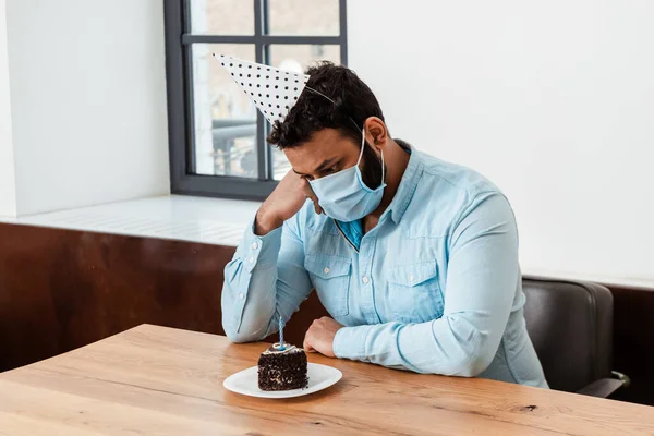 Hombre afroamericano en gorra de fiesta y máscara médica celebrando cumpleaños solo y mirando pastel de cumpleaños en el plato - foto de stock