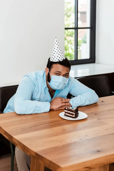 Африканский американец в шапочке для вечеринок и медицинской маске, празднующий день рождения в одиночестве рядом с вкусным праздничным тортом — стоковое фото
