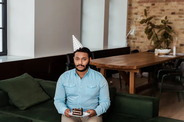 Hombre afro-americano trastornado en gorra de fiesta celebrando cumpleaños y sosteniendo plato con pastel de cumpleaños - foto de stock