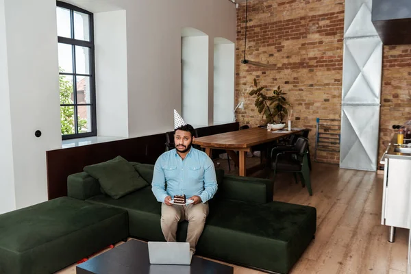 Frustrated african american man in party cap celebrating birthday and holding plate with birthday cake near laptop — Stock Photo