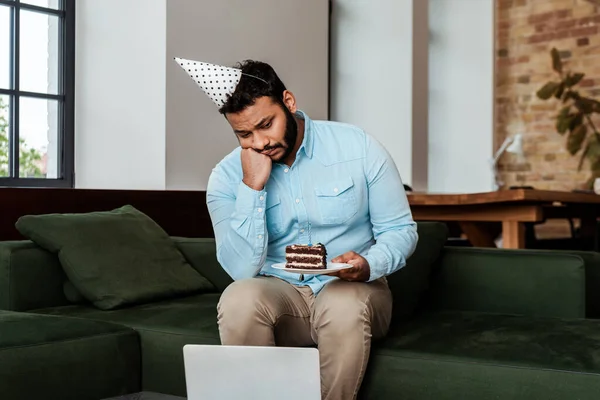 Frustrated african american man in party cap celebrating birthday and holding plate with birthday cake while looking at laptop — Stock Photo