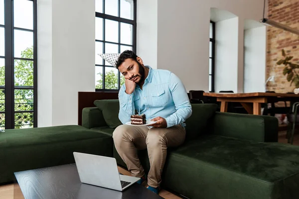 Uomo africano americano frustrato in tappo partito in possesso di piatto con torta di compleanno vicino al computer portatile — Foto stock