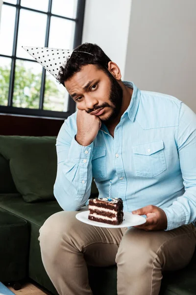 Frustrated african american man in party cap holding plate with birthday cake — Stock Photo