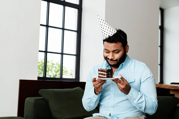 Недовольный африканский американец в шапочке для вечеринок держит тарелку с праздничным тортом — стоковое фото