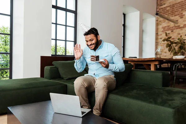 Hombre afroamericano feliz en la tapa del partido sosteniendo pastel de cumpleaños y saludando la mano mientras que tiene videollamada - foto de stock