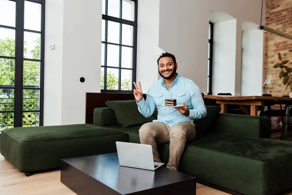 Glücklich afrikanisch-amerikanischer Mann in Partymütze hält Geburtstagstorte und zeigt Frieden singen in der Nähe von Laptop — Stockfoto