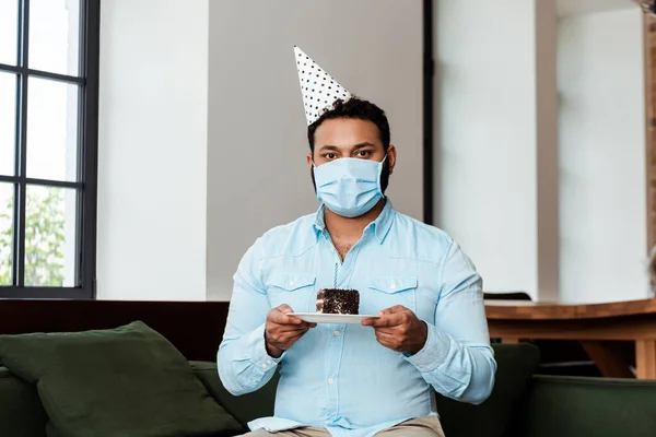 Homem americano africano no tampão do partido e na máscara médica que prende a placa com bolo de aniversário — Fotografia de Stock