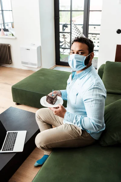 African american man in party cap and medical mask holding with plate birthday cake near laptop in living room — Stock Photo