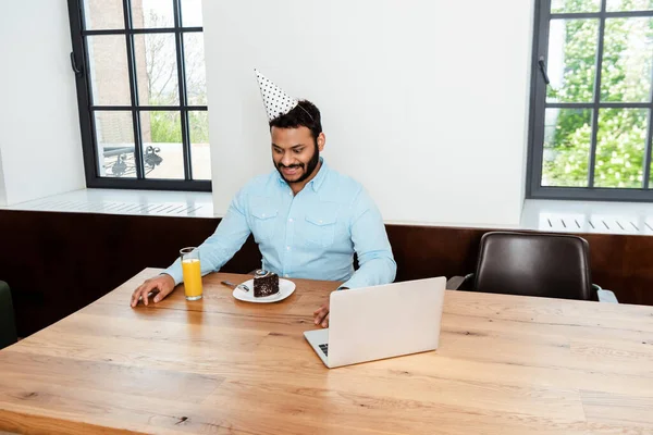 Heureux homme afro-américain en chapeau de fête en regardant gâteau d'anniversaire savoureux près d'un ordinateur portable et un verre de jus d'orange — Photo de stock