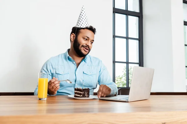 Homme afro-américain en chapeau de fête regardant ordinateur portable tout en ayant appel vidéo près gâteau d'anniversaire savoureux et verre de jus d'orange — Photo de stock