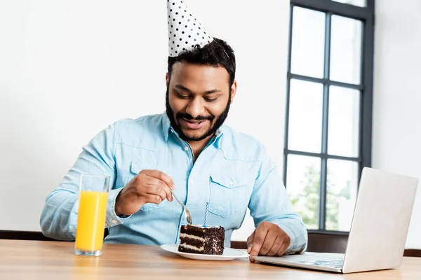 Fröhlicher afrikanisch-amerikanischer Mann mit Partymütze blickt auf leckeren Geburtstagskuchen neben Laptop und einem Glas Orangensaft — Stockfoto