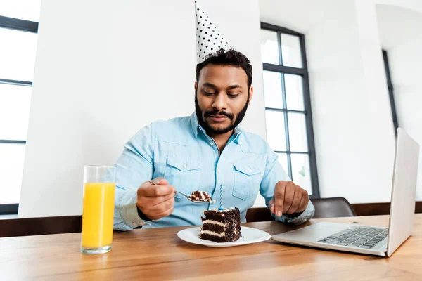 Homme afro-américain en chapeau de fête en regardant gâteau d'anniversaire savoureux près d'un ordinateur portable et un verre de jus d'orange — Photo de stock