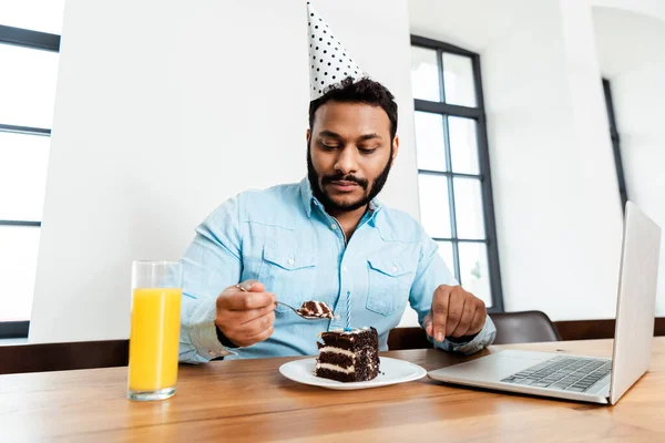 Afrikanisch-amerikanischer Mann mit Partymütze blickt auf leckeren Geburtstagskuchen neben Laptop und einem Glas Orangensaft — Stockfoto