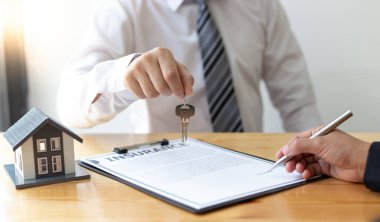 Emlakçı müşteriye gönderilecek anahtarları elinde tutuyor ve ev satışı anlaşmasını ve ev sigortasını müşteriye, ev satışları ve ev sigortası kavramına imzalama şartlarını sunuyor..