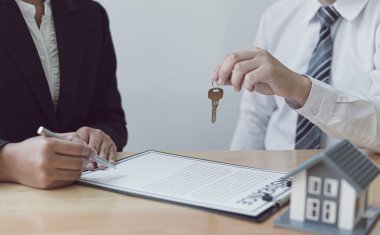 Emlakçı müşteriye gönderilecek anahtarları elinde tutuyor ve ev satışı anlaşmasını ve ev sigortasını müşteriye, ev satışları ve ev sigortası kavramına imzalama şartlarını sunuyor..