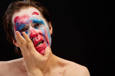 Genç bir adam tek eliyle yüzüne palyaço makyajı sürer, çıplak omuzlarıyla, duyguları ifade eder, siyah bir arka planda.